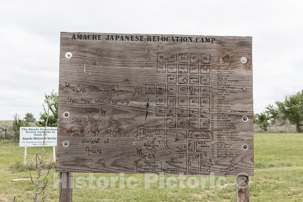 Photograph- An etched wooden sign"Amache Japanese Relocation Camp," complete with outlines of buildings, one of the only remnants, save for a few cement foundations a little nearby graveyard