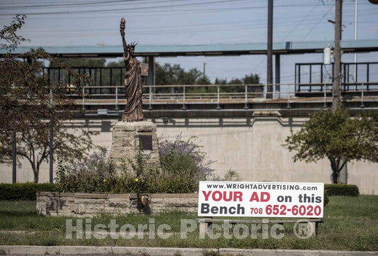 Photo- A garish Bench Advertisement diminishes The Grandeur of a Nearby, Scaled-Down Model of New York Harbor's Statue of Liberty, Standing Near a Train Overpass in Gary, Indiana